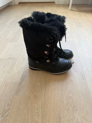 Svarta moon boots med fuskpäls - Säljer ett par svarta moon boots med snörning och detaljer i metall. Bootsens insida och kant är fodrade med mjuk, fluffig fuskpäls för extra värme. Yttermaterialet är en mix av textil och syntet, perfekta för kalla vinterdagar. Finns två par i storlek 35 respektive 36 