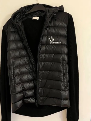 Moncler  - Moncler stickad dunjacka med huva i svart. Kombination av stickade ärmar och rygg med lättviktsdun framtill ger en stilren och exklusiv look. Två dragkedjefickor framtill och genomgående dragkedja. Jackan är i mycket fint skick utan hål eller större slitage. Perfekt som höst-, vår- eller mild vinterjacka.