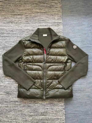 Olivgrön cardigan från Moncler - Säljer en olivgrön pufferjacka från Moncler med stickade ärmar och krage. Jackan har quiltad front i glansigt material, dragkedja och en Moncler-logga på ärmen. Känn er fria att fråga om hur må ga bilder ni vill. ❗️❗️den är riktig❗️❗️notera även slitage på sista bilden❗️❗️jackan är i storlek XL men passar mig mellen M och L.