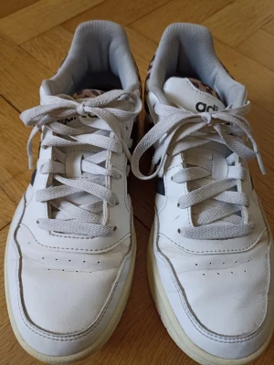 Vita Adidas sneakers med leoparddetaljer - Snygga vita sneakers från Adidas med leopardmönstrade detaljer på hälen. Skorna har klassisk låg profil, vita snören och robust sula. Tillverkade i läder och textil för en fräsch och trendig look. Perfekta för dig som vill sticka ut lite extra.