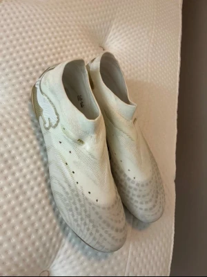 Vita och guldiga fotbollsskor Puma - Säljer ett par stilrena fotbollsskor från Puma i vitt med subtila mönster och guldfärgade dobbar. Skorna har en strömlinjeformad design utan snörning och är tillverkade i syntetmaterial för bra grepp och känsla på planen.