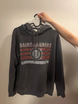 Mörkgrå Saint Laurent hoodie - Mörkgrå hoodie från Saint Laurent med tryck i vitt och rött framtill. Klassisk känguruficka, huva med dragsko och ribbade muddar. Snygg streetstil med tydlig logga och grafiska ränder på bröstet. Strl M men jag skulle säga Small