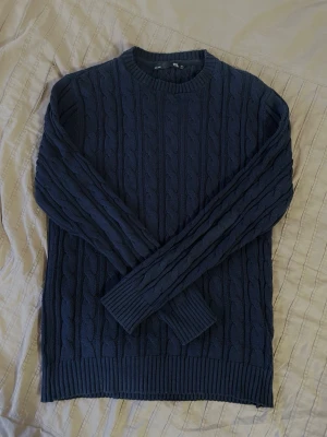 Marinblå kabelstickad tröja - Säljer en marinblå kabelstickad tröja med rund halsringning och ribbade muddar vid ärmslut och nederkant, vilket ger en klassisk look. Perfekt att ha över en skjorta! Tröjan har dock krympt lite pga. tvätt, så ha i åtanke att den kan sitta lite mindre än M. Den är gjord av 100% bomull.  Axelbredd: 45 cm.  Längd: 71 cm.