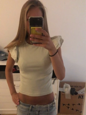 Ljusvit topp med volangärmar - Säljer en ljusvit croppad topp med volangdetaljer på ärmarna. Toppen är ärmlös och har en tight passform som framhäver midjan. Perfekt till jeans eller kjol för en fräsch look. Är i storlek 146/152 men sitter som xxs!