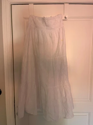 Vit långkjol med broderade detaljer - Luftig vit långkjol med broderade blommönster och smock i midjan. Kjolen har en volang och är tillverkad i ett lätt bomullstyg som passar perfekt till varmare dagar.