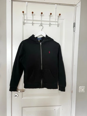 Svart hoodie från Polo Ralph Lauren - Säljer en svart hoodie från Polo Ralph Lauren med dragkedja framtill och två fickor. Klassisk huva och den ikoniska röda loggan broderad på bröstet. Perfekt för en avslappnad och stilren look. Tillverkad i mjukt bomullsmaterial.