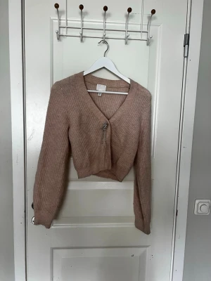 Beige stickad kofta med rosett - Supersöt beige stickad kofta med v-ringning och lång ärm. Koftan är croppad och har en glittrig rosett-detalj framtill som stängning. Perfekt att styla över ett linne eller klänning för en trendig look.