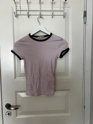 Ljusrosa t-shirt från Bershka - Säljer en ljusrosa t-shirt från Bershka med svarta kontrastkanter vid hals och ärmslut. Modellen är kortärmad och har en tight passform.
