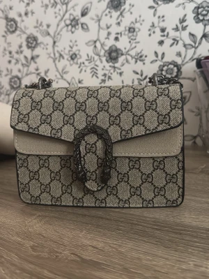  GG Supreme handväska - Säljer en snygg  Dionysus handväska i beige och grått GG Supreme mönster. Väskan har silverfärgad kedjerem och ett dekorativt spänne framtill. Materialet är canvas med detaljer i mocka. Perfekt för dig som gillar lyxiga accessoarer med ikonisk design.