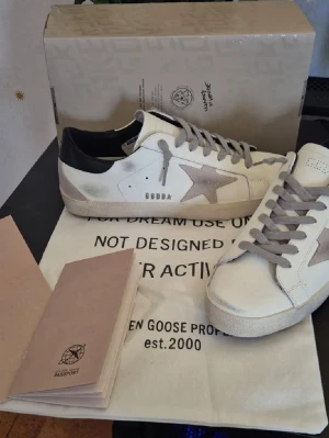 Golden Goose vita sneakers med stjärna - Golden Goose sneakers i vitt läder med grå mockadetaljer och ikonisk stjärna på sidan. Skorna har grå skosnören, svart hälparti och en vintage-inspirerad sula. Perfekta för dig som gillar en avslappnad men trendig stil.