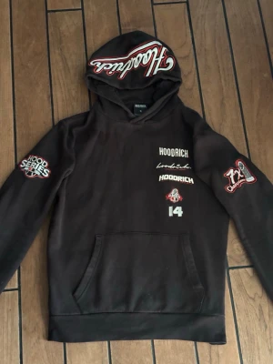 Hoodrich set  - Svart set från Hoodrich med flera vita och röda tryck på bröstet, ärmarna, huvan och ryggen. Och på byxorna finns det tryck vid vänstra låret och på bakfickan. Materialet är mjukt och bekvämt, perfekt för en streetwear-look. 