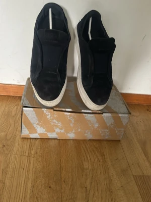 Svarta slip-on sneakers i mocka - Stilrena svarta slip-on sneakers med ovandel i mocka och vit sula. Skorna har en minimalistisk design utan synliga snören och en låg siluett som passar till många olika outfits. Perfekta för dig som gillar enkel och clean stil.
