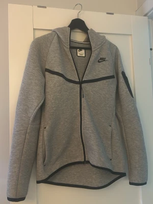 Grå Nike hoodie med dragkedja - Snygg grå hoodie från Nike med hel dragkedja och svarta detaljer. Hoodien har huva, två fickor framtill och en svart Nike-logga på bröstet. 