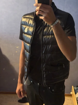 Svart dunväst från Moncler - Snygg svart dunväst från Moncler med glansig finish och klassisk logga på bröstet. Västen har ståkrage, dragkedja framtill och två fickor med dragkedja. Perfekt för lager-på-lager och ger en clean look.