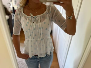 Vit blus med blått mönster Hollister - Söt vit blus från Hollister med ljusblått blommigt mönster och knyt i halsen. Blusen har trekvartsärm, volang nedtill och är tillverkad i ett tunt, luftigt material. Perfekt för en fräsch och somrig look.