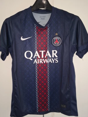 PSG tröja O. Dembélé #10 - Mörkblå PSG matchtröja från Nike med O. Dembélé och nummer 10 på ryggen. Tröjan har Qatar Airways-tryck på bröstet, PSG-logga och vit Nike-swoosh. Röd och vit grafisk rand i mitten och korta ärmar. Perfekt för fotbollsfans!