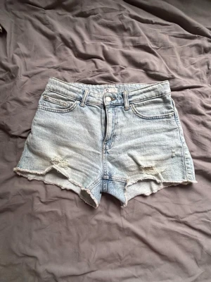 Ljusblå denimshorts med råa kanter - Säljer ett par ljusblå denimshorts med fransiga, råa kanter och slitna detaljer framtill. Klassisk femficksmodell med knapp och dragkedja. Perfekta för varma dagar och en avslappnad stil.