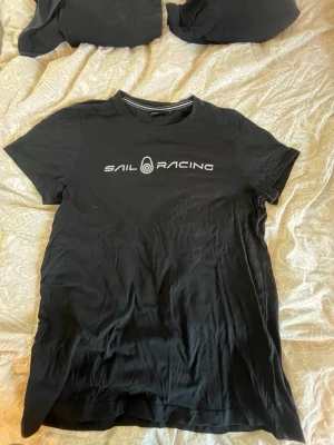 Svart Sail Racing t-shirt - Svart t-shirt från Sail Racing med vit logga tryckt på bröstet. Klassisk rund hals och korta ärmar. Tillverkad i mjuk bomull som känns skön mot huden. Perfekt för en avslappnad och sportig stil.