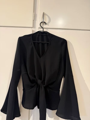 Svart blus med knyt och vida ärmar - Snygg svart blus med v-ringning och knyt i midjan. Blusen har långa, vida ärmar och är gjord i ett mjukt, glansigt tyg som ger en elegant känsla. Perfekt att styla med jeans eller kjol för en trendig look.