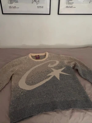 Corteiz sweater / M - Säljer en gråmelerad stickad tröja med stor vit C och stjärna på framsidan. Tröjan har rund halsringning, ribbade muddar och nederkant samt en liten broderad detalj på ärmen. Materialet ser mjukt och fluffigt ut, perfekt för kyliga dagar. Endast provad