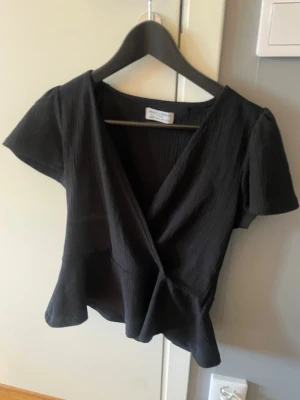 Svart omlottblus Holly & Whyt by Lindex  - Snygg svart omlottblus från Lindex med korta ärmar och lätt struktur i tyget. Blusen har en djup v-ringning och markerad midja som ger en feminin siluett. Perfekt att matcha med både jeans och kjol.