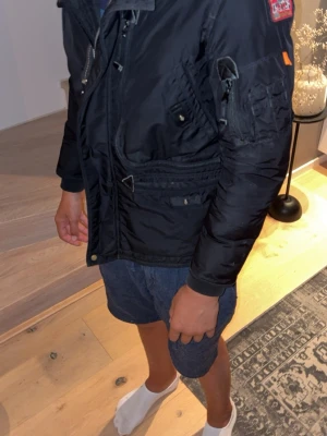 Parajumper vinterjacka  - Säljer en svart jacka med flera fickor, dragkedja och tryckknappar framtill. Jackan har en patch på ärmen och detaljer som ger en cool look. Materialet ser ut att vara syntetiskt och jackan har ribbade muddar vid ärmsluten. Perfekt för dig som gillar streetwear.