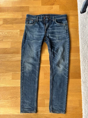 Blå  jeans från Nudie Jeans - Säljer ett par klassiska blå slim fit jeans från Nudie Jeans med snygg slitning och orangea sömmar. Jeansen har fem fickor, knappgylf och en bekväm passform. Perfekta till vardags för en avslappnad men stilren look. W31 L32