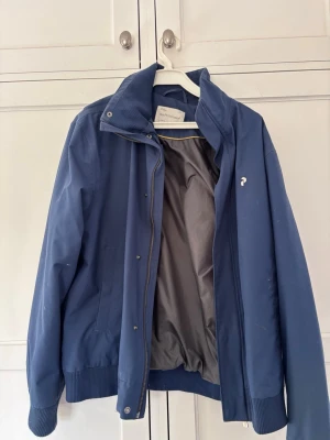 Blå vindjacka med hög krage - Snygg blå vindjacka med hög krage och ribbade muddar. Jackan har dragkedja och tryckknappar framtill samt en liten vit logga på bröstet. Insidan är fodrad i grått och materialet känns lätt och vindtätt. Billigare vid snabbare affär 