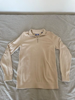 Beige stickad tröja med halv dragkedja - Snygg beige stickad tröja med halv dragkedja framtill och hög krage. Tröjan har långa ärmar och ribbade muddar vid ärmslut och nederkant. Perfekt för lager på lager och enkel att matcha med olika outfits.