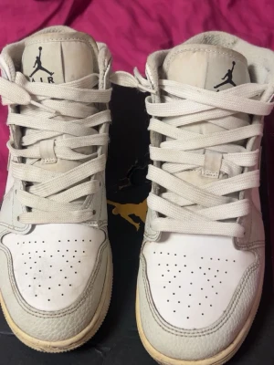 Air Jordan 1 Mid sneakers - Säljer ett par vita Air Jordan 1 Mid sneakers med ljusgrå camo detaljer och klassisk Jumpman-logga på plösen. Skorna har en ikonisk siluett, perforerad tåbox och är tillverkade i läder med textildetaljer. Perfekta för dig som gillar streetwear och snygg design. Obs original skokartong följer inte med