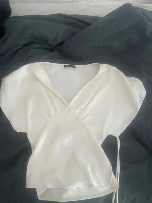 Vit omlottblus i satin - Snygg vit omlottblus i glansigt satinmaterial med korta ärmar och knytband i midjan. Blusen har en djup v-ringning och en lätt figurnära passform som ger en elegant look. Perfekt att styla med jeans eller kjol för en trendig outfit.