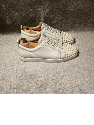  Louboutin - Säljer ett par vita sneakers från Christian Louboutin i läder med coola vita nitar på tån. Skorna har klassisk låg profil, vita snören och en stilren design som sticker ut. Perfekta för dig som vill ha något extra till din outfit.