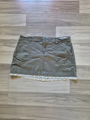Grön kortkjol med spetskant - Snygg grön kortkjol i bomull med två stora fickor fram och dekorativ vit spetskant längst ner. Kjolen har bälteshällor.. Perfekt för en avslappnad och trendig look.