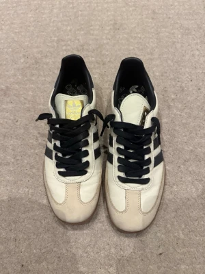 Adidas Samba vita och svarta  - Klassiska Adidas Samba sneakers i vitt läder med svarta ränder och svart snörning. Skorna har en brun gummisula och gulddetaljer på plösen. Perfekta för en sportig och stilren look. Låg modell som passar till det mesta.
