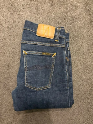 Mörkblå smala jeans Nudie Jeans - Snygga mörkblå jeans från Nudie Jeans med smal passform och klassiska gula sömmar. Jeansen har två bakfickor med dekorativa detaljer och ett läderpatch med logga i midjan. Perfekta till en avslappnad och trendig stil. Storleken är W30/L32. Pris kan diskuteras. Skriv vid minsta fråga. 