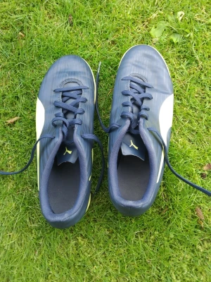 Blåa fotbollsskor från Puma - Snygga blåa inomhusskor från Puma med vita detaljer och klassisk snörning. Skorna har en låg profil och är tillverkade i syntetmaterial som ger bra fäste i marken och komfort på idrotten Perfekta för dig som vill ha stilrena och funktionella skor till match eller träning. Pris kan diskuteras 
