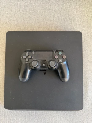 PlayStation 4 Slim med handkontroll - Säljer en svart PlayStation 4 Slim med tillhörande svart DualShock 4-handkontroll. Konsolen har en stilren, fyrkantig design och är tillverkad i plast. Perfekt för gaming och filmkvällar.