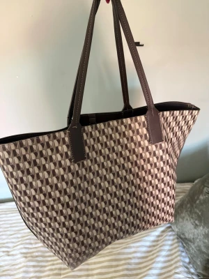 Väska från Mango - Stilren shoppingväska i brunt och beige med grafiskt mönster. Väskan har långa, bruna handtag i konstläder och rymlig insida. Perfekt för dig som vill ha plats för allt och gillar en trendig look. Materialet känns tåligt och väskan har en klassisk, fyrkantig form. Handtaget är lite slitet längst upp🤎
