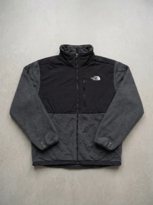 Grå fleecejacka från The North Face - Snygg grå fleecejacka från The North Face med svarta partier över axlar och bröst. Jackan har hög krage, dragkedja framtill och två sidofickor med dragkedja. Perfekt för lager-på-lager och outdoor-vibe. Storlek: L
