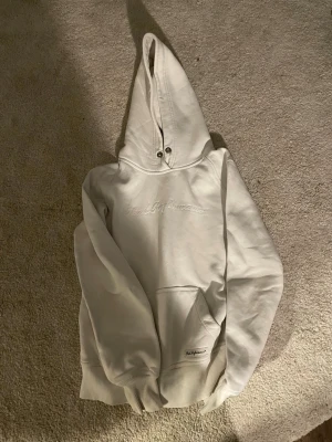 Vit hoodie från Peak Performance - Säljer en vit hoodie från Peak Performance med broderad logga på bröstet och stor känguruficka framtill. Hoodien har ribbade muddar vid ärmslut och nederkant samt en klassisk huva med metallöljetter. Perfekt för en chill och stilren look.