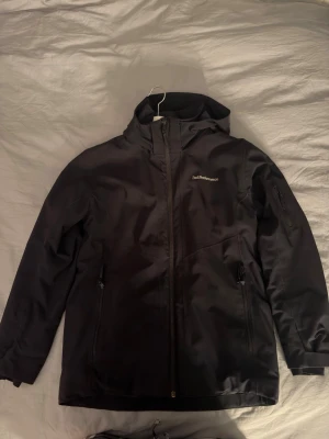 Svart vindjacka från Peak Performance - Snygg svart vindjacka från Peak Performance med huva och dragkedja framtill. Jackan har två sidofickor med dragkedja och en extra ficka på ärmen. Tillverkad i ett slitstarkt material som står emot vind och lätt regn. Perfekt för utomhusaktiviteter. Storlek 160 men passar xs