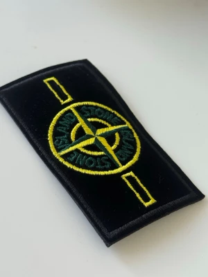 Stone Island badge - Säljer ett klassiskt Stone Island tygmärke i svart med broderad gul och grön kompasslogga. Märket är rektangulärt och har detaljer i gult och grönt, perfekt att sy eller fästa på jackor, väskor eller kepsar.