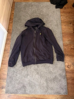 Mörkblå hoodie från Lyle & Scott - Snygg mörkblå hoodie från Lyle & Scott med dragkedja framtill och klassisk gul logga på bröstet. Tröjan har huva med snörning och två fickor framtill. Tillverkad i mjukt bomullsmaterial som är skönt att bära.