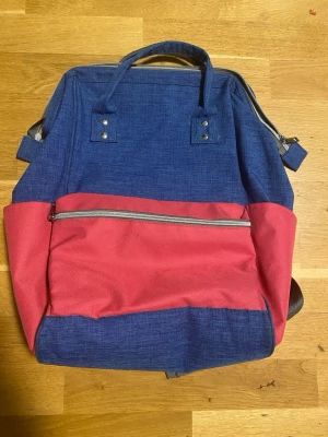 Blå och röd ryggsäck med dragkedja - Snygg ryggsäck i blått och rött tyg med en stor ytterficka med dragkedja framtill. Väskan har två handtag upptill och justerbara axelband. Perfekt för skolan eller utflykter, och har en modern, boxig form. Materialet är slitstark textil.