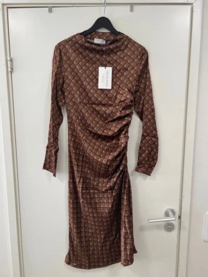 Brun mönstrad långklänning - Säljer en brun långklänning med rutigt mönster och snygg rynkning på sidan. Klänningen har långa ärmar och en figurnära passform. Materialet känns glansigt och mjukt, perfekt för dig som vill sticka ut med en unik look.