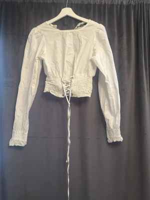 Vit blus med snörning i midjan - Trendig vit blus med långa ärmar och smockdetaljer vid ärmslut och midja. Blusen har en snygg snörning framtill som ger en cool och feminin siluett. Perfekt att styla med både jeans och kjol för en chic look.