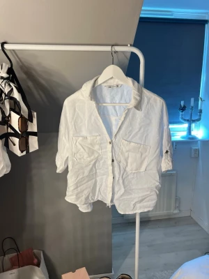 Vit skjorta med fickor och knappar - Vit skjorta i avslappnad passform med två stora bröstfickor och knäppning framtill. Skjortan har krage och korta ärmar, perfekt för en stilren och enkel look. Tillverkad i ett lätt material som känns svalt.