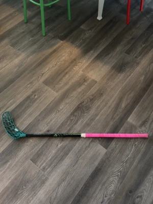 Innebandyklubba med turkost blad - Säljer en innebandyklubba med svart skaft, turkost plastblad och rosa grepp. Klubban har en modern design och är perfekt för dig som gillar innebandy. Greppet är lindat med rosa tejp för extra stilpoäng. Den är 96 cm och right blad