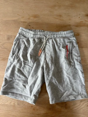 Grå mjukisshorts med snörning - Snygga grå mjukisshorts med elastisk midja och snörning framtill. Shortsen har två sidofickor och en bakficka med liten röd detalj. Perfekta för chill dagar eller träning. Tillverkade i mjukt bomullsmaterial för maximal komfort.