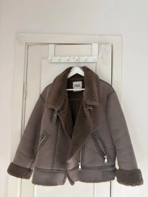 Brun teddyjacka från ZARA - Säljer en brun teddyjacka från ZARA med bred krage och dragkedjor på fickorna. Jackan har mjukt, fluffigt teddyfoder och detaljer i samma färg på ärmslut och krage. Perfekt för kalla dagar och riktigt mysig att bära.